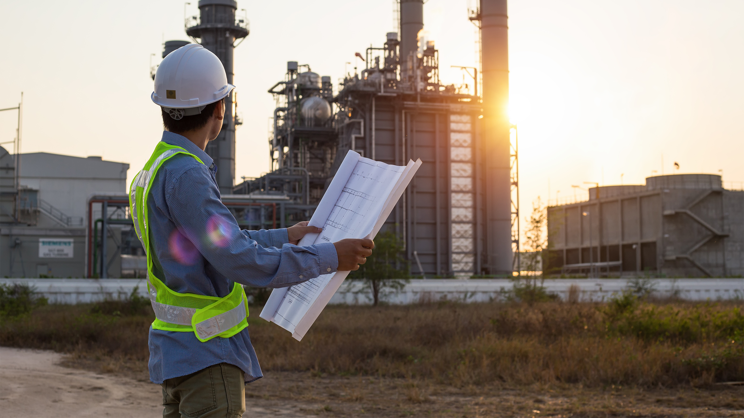 Engineer in safety vest reviews blueprints of a large power plant at sunset.