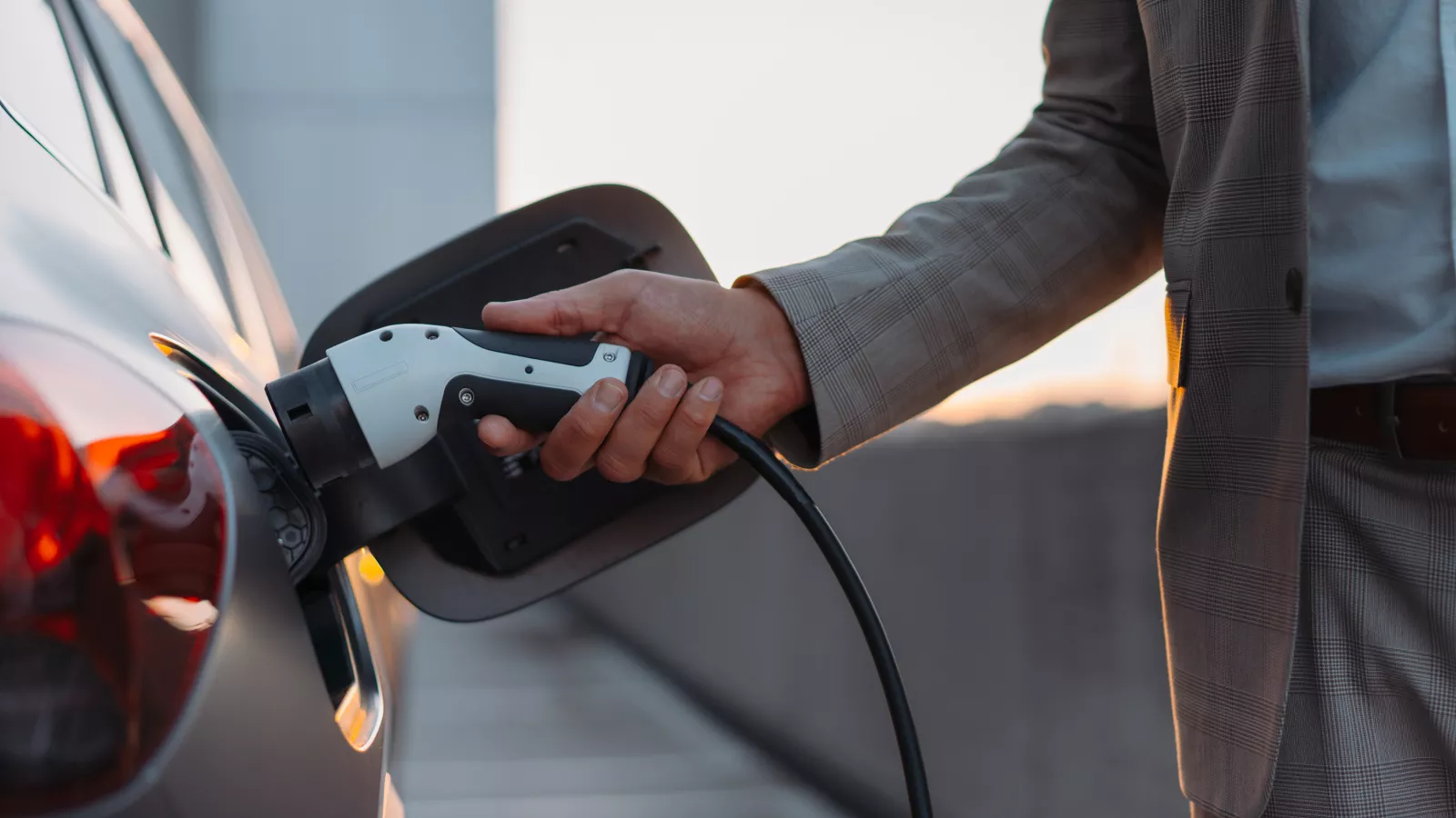A man in a stylish checkered suit is charging his electric vehicle at a charging station. The focus is on his hand holding the charging cable and connecting it to the car. The scene takes place on a parking deck at sunset.
