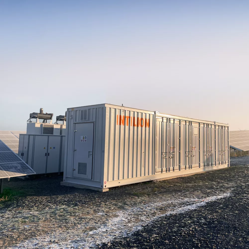 Large, light gray, modular containerized power equipment, labeled INTILION, situated amongst solar panels.
