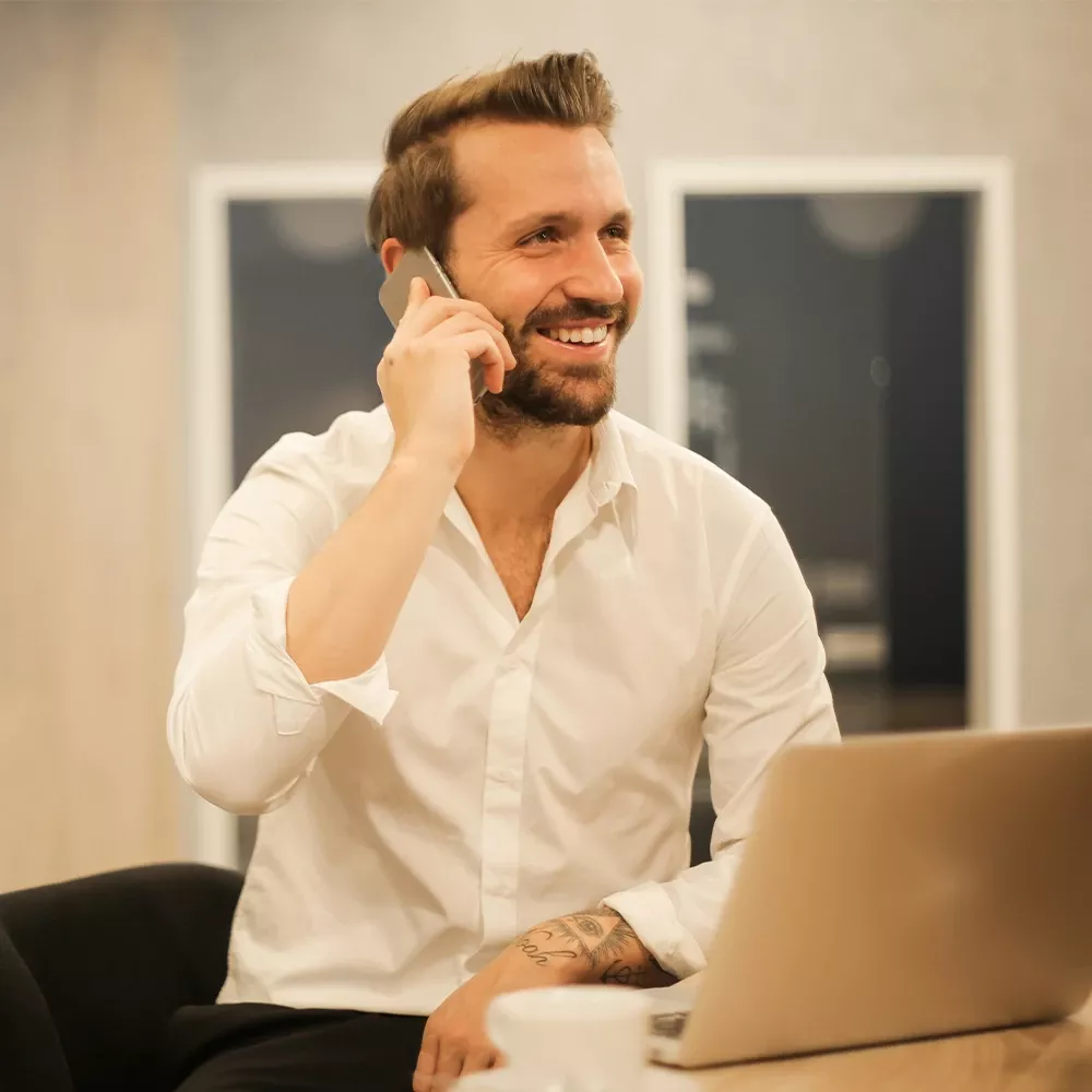 Man in a white shirt smiling while talking on the phone at a desk with a laptop.